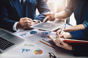 Three people in a business meeting review financial data on a tablet, with charts and graphs on the table.best financial service for middle class families.