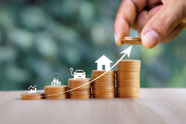 A hand adds a coin to a growing stack, with icons for a house, car, and factory along a line of coins, representing the growth of various investments.best financial services for middle class families