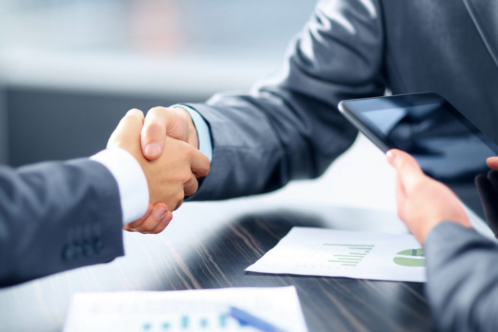 Two business people shaking hands over a table with a tablet and documents, signifying a deal or agreement.