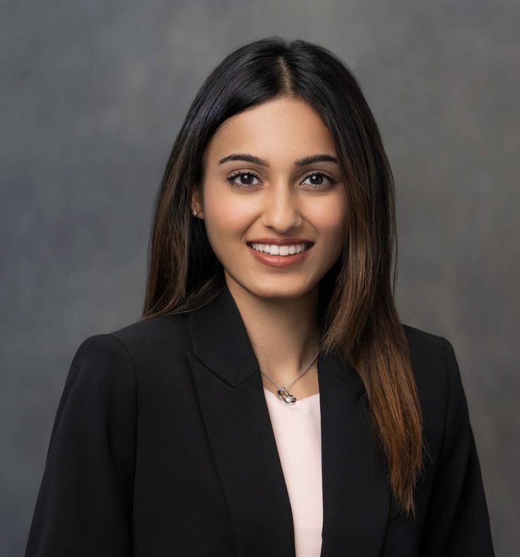 A headshot of a smiling young woman with long dark hair, wearing a black blazer.